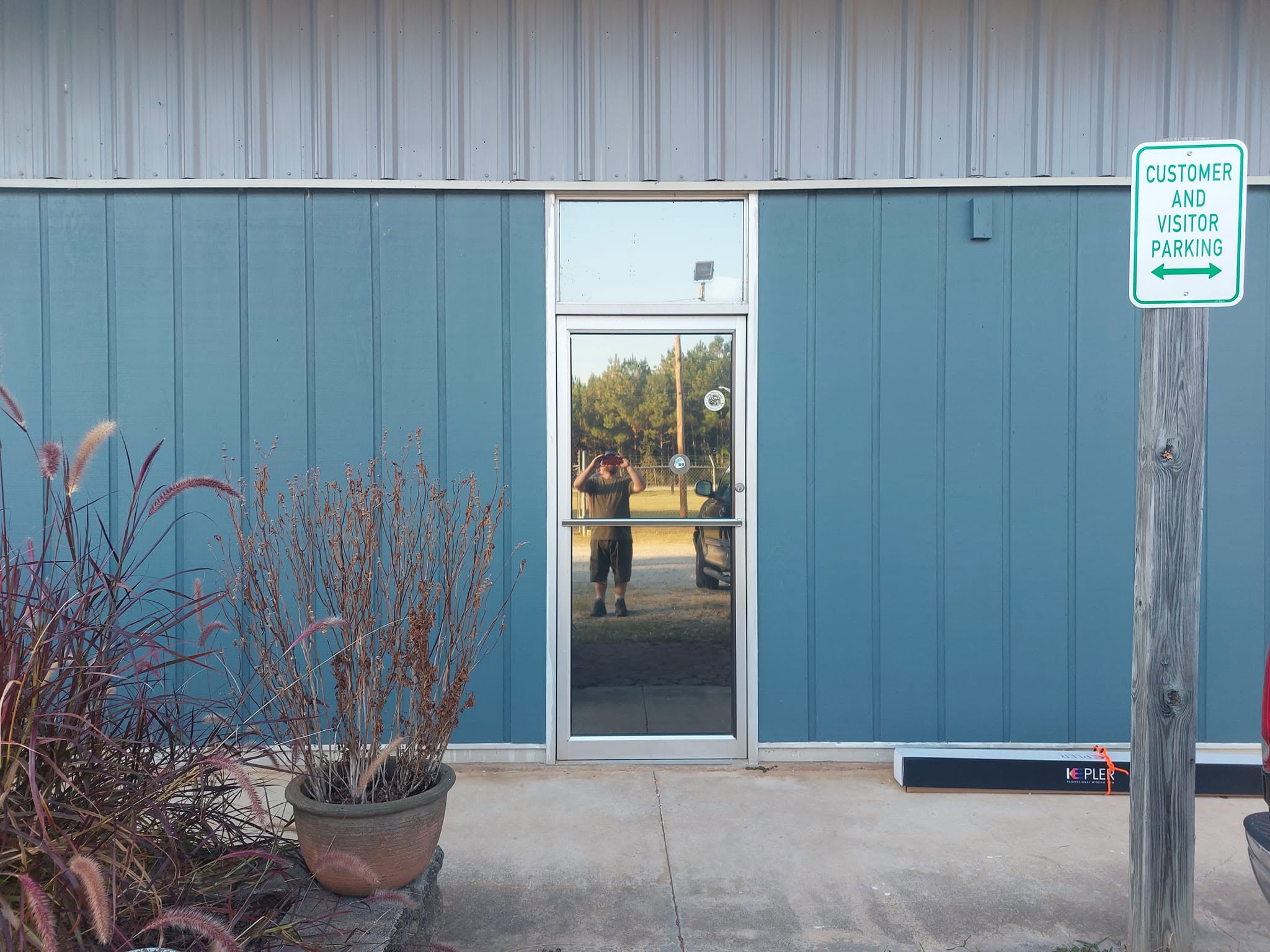Storefront entrance with glass door, blue metal siding, potted ornamental grass, and customer parking sign