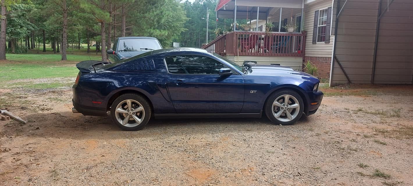 Dark blue Ford Mustang GT parked on gravel driveway beside residential home