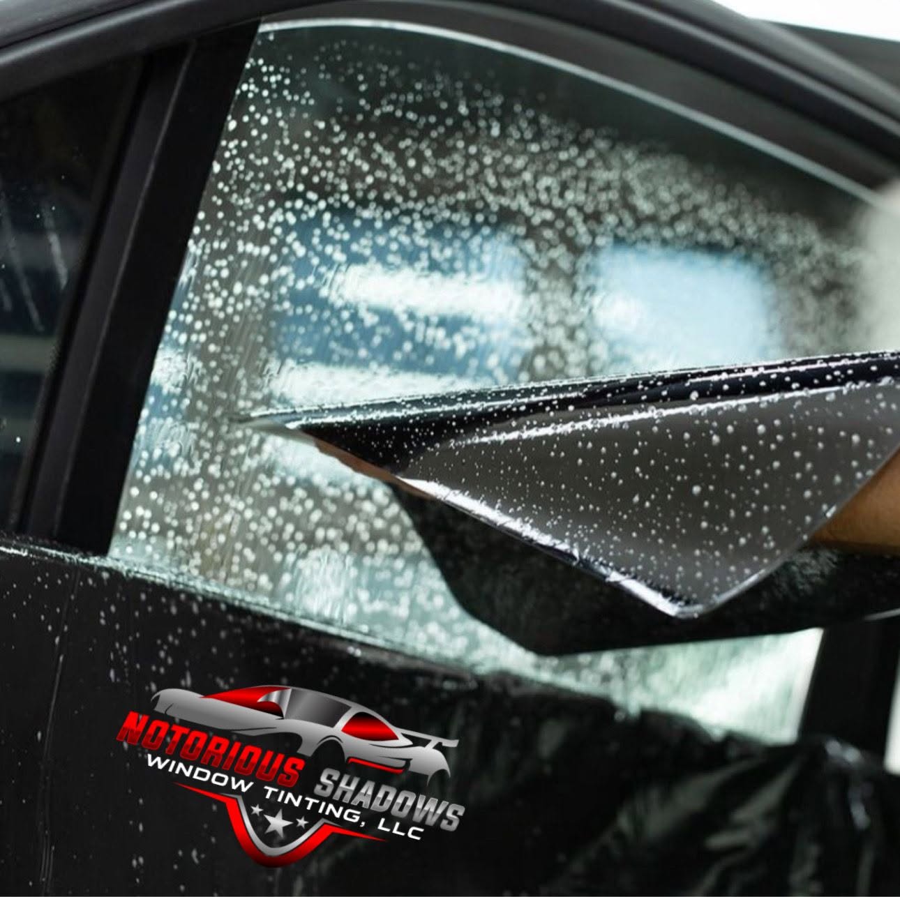 Car window covered in water droplets during washing with squeegee tool and Notorious Shadows logo visible