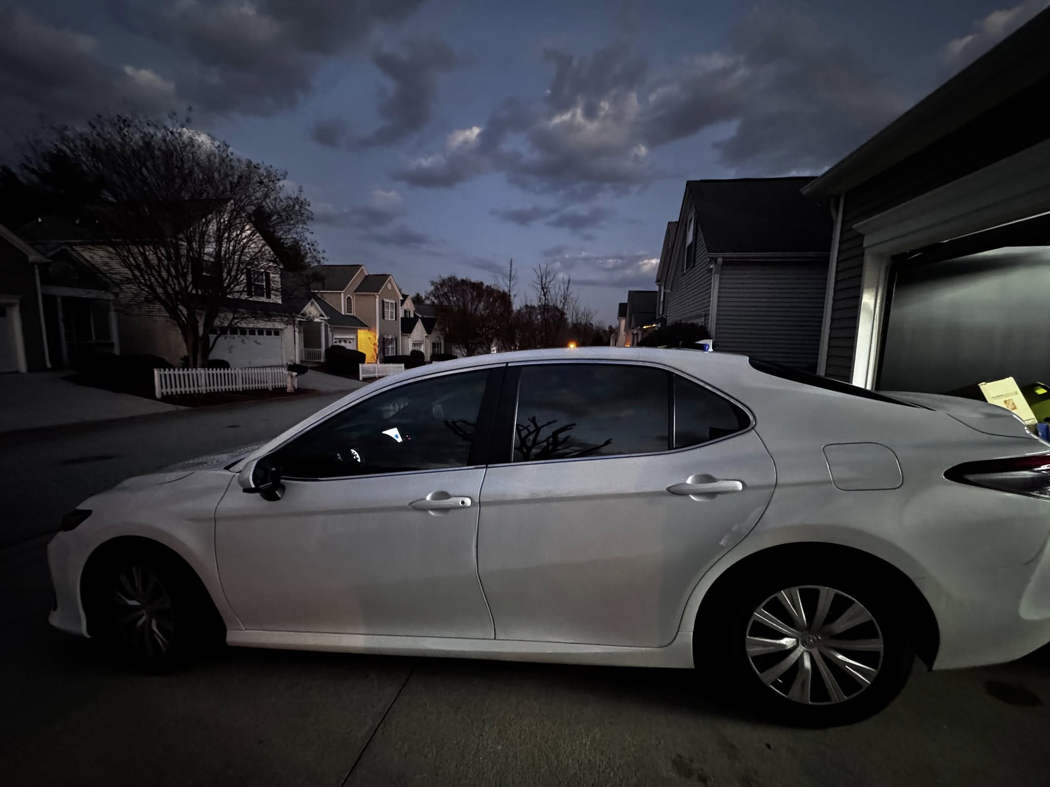 White sedan parked in suburban driveway at dusk with houses and bare trees in background